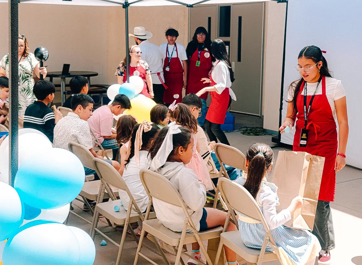 Niños sentados bajo toldo con voluntarios en evento infantil de Vino Nuevo.